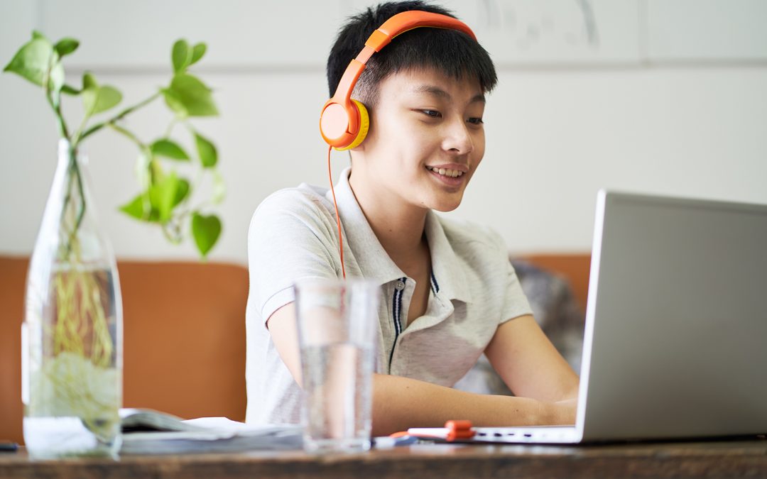  A teenage boy wearing orange headphones is smiling while studying at a table with a laptop. A glass of water and a plant in a clear vase are also visible on the table.