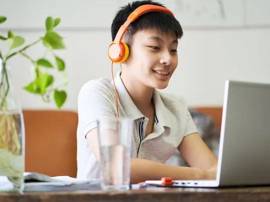 A teenage boy wearing orange headphones is smiling while studying at a table with a laptop. A glass of water and a plant in a clear vase are also visible on the table.