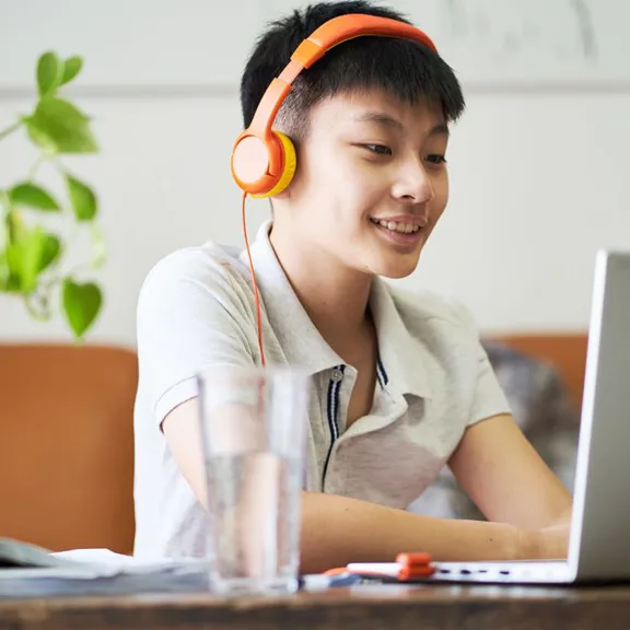 A teenage boy wearing orange headphones is smiling while studying at a table with a laptop. A glass of water and a plant in a clear vase are also visible on the table.