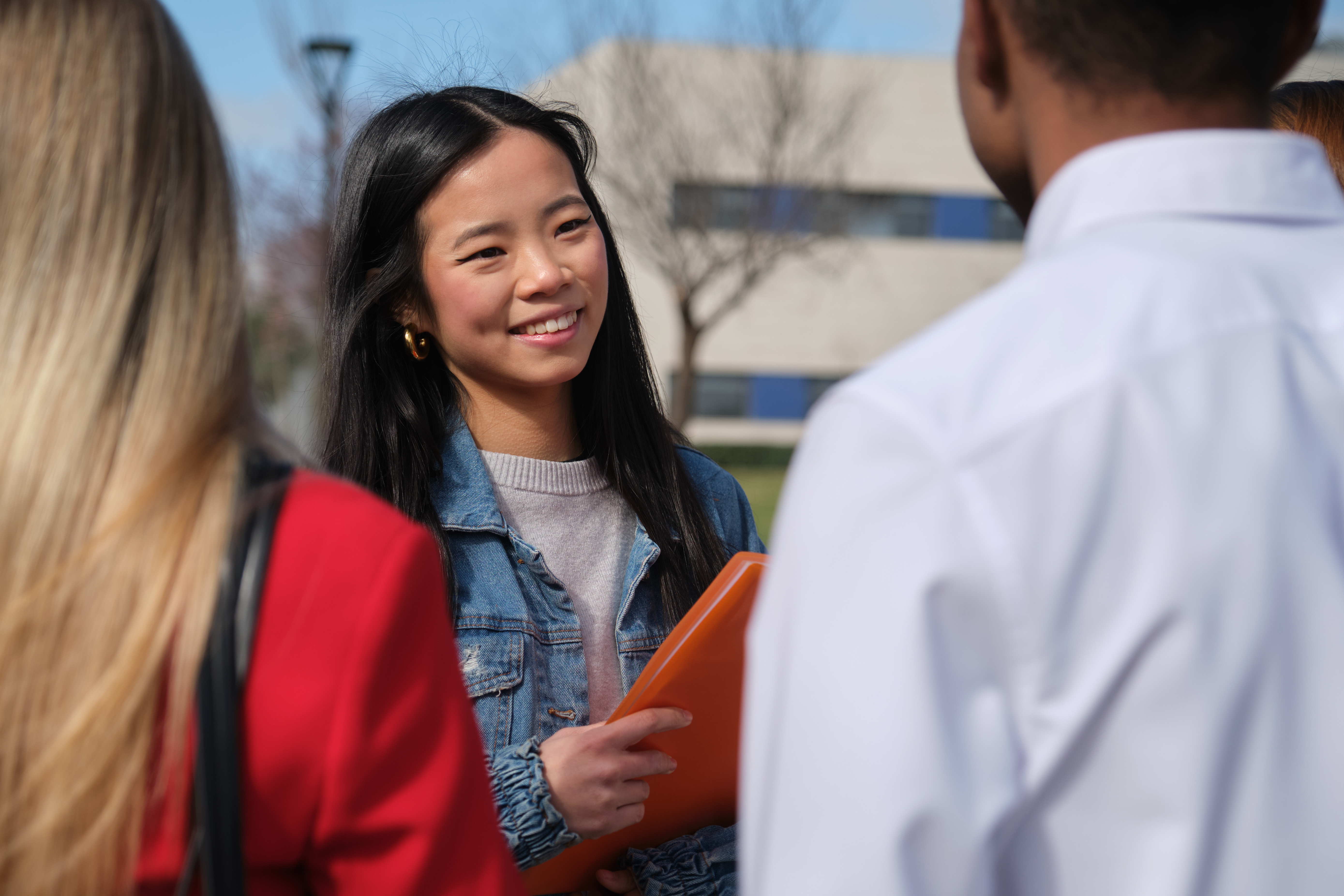 A young woman in a denim jacket smiles brightly while holding an orange folder and discussing with two classmates on a sunny school campus.