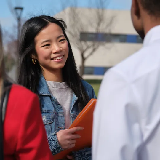 A young woman in a denim jacket smiles brightly while holding an orange folder and discussing with two classmates on a sunny school campus.