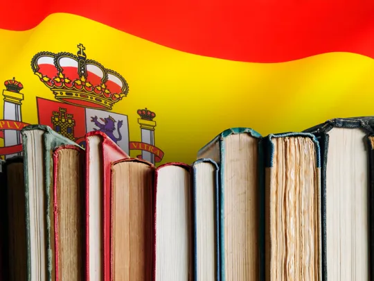 A close-up of a row of books against a background of the Spanish flag and coat of arms, symbolizing the study of the Spanish language.