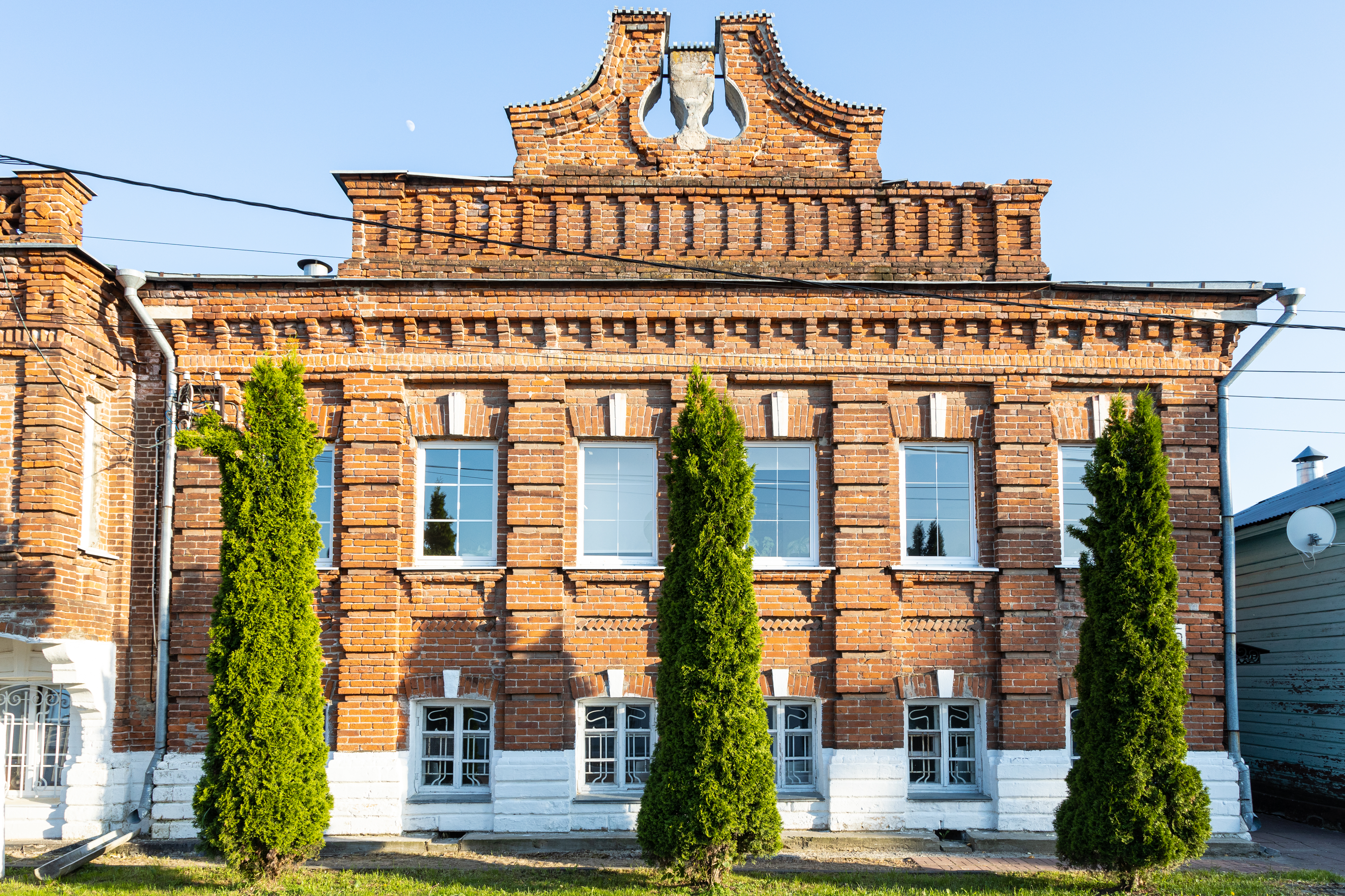 The exterior of an old, ornate two-story red brick school building with tall, green evergreen trees lined up in front of the windows.