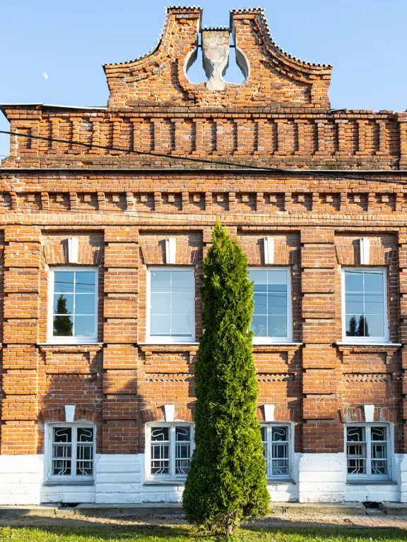The exterior of an old, ornate two-story red brick school building with tall, green evergreen trees lined up in front of the windows.