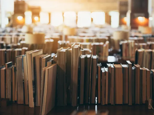 A warm, inviting photo inside a library, showing rows of old books standing upright on tables with sunlight streaming in the background.