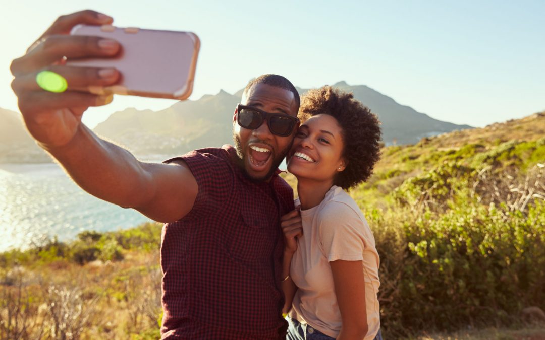A happy couple taking a selfie with a smartphone while standing on a hill overlooking a body of water and mountains. The man in the foreground is holding the phone and has a wide-open smile, while the woman next to him is also smiling and leaning in.