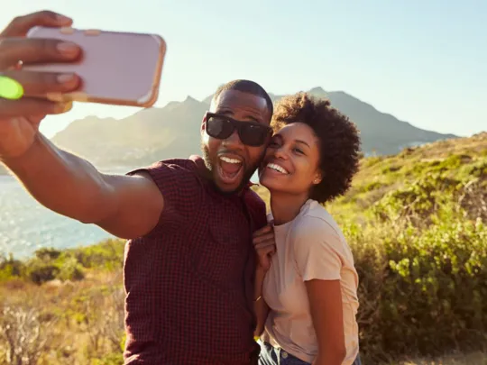 A happy couple taking a selfie with a smartphone while standing on a hill overlooking a body of water and mountains. The man in the foreground is holding the phone and has a wide-open smile, while the woman next to him is also smiling and leaning in.