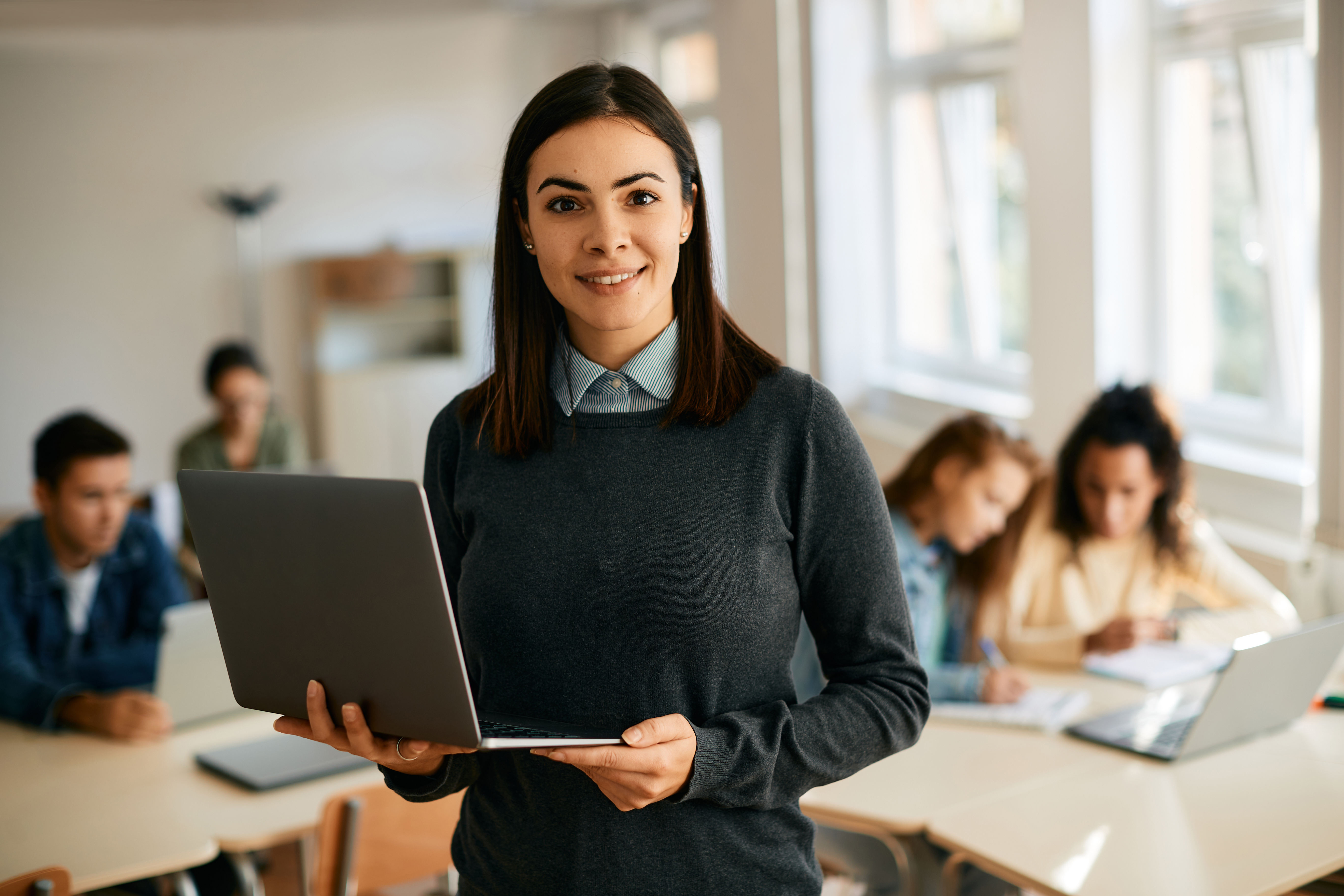 A smiling female teacher in a gray sweater holding a laptop, standing in the middle of a classroom with other students working at desks behind her.