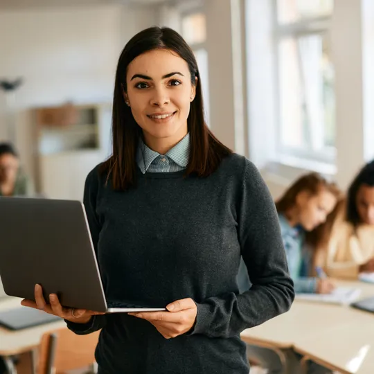 A smiling female teacher in a gray sweater holding a laptop, standing in the middle of a classroom with other students working at desks behind her.