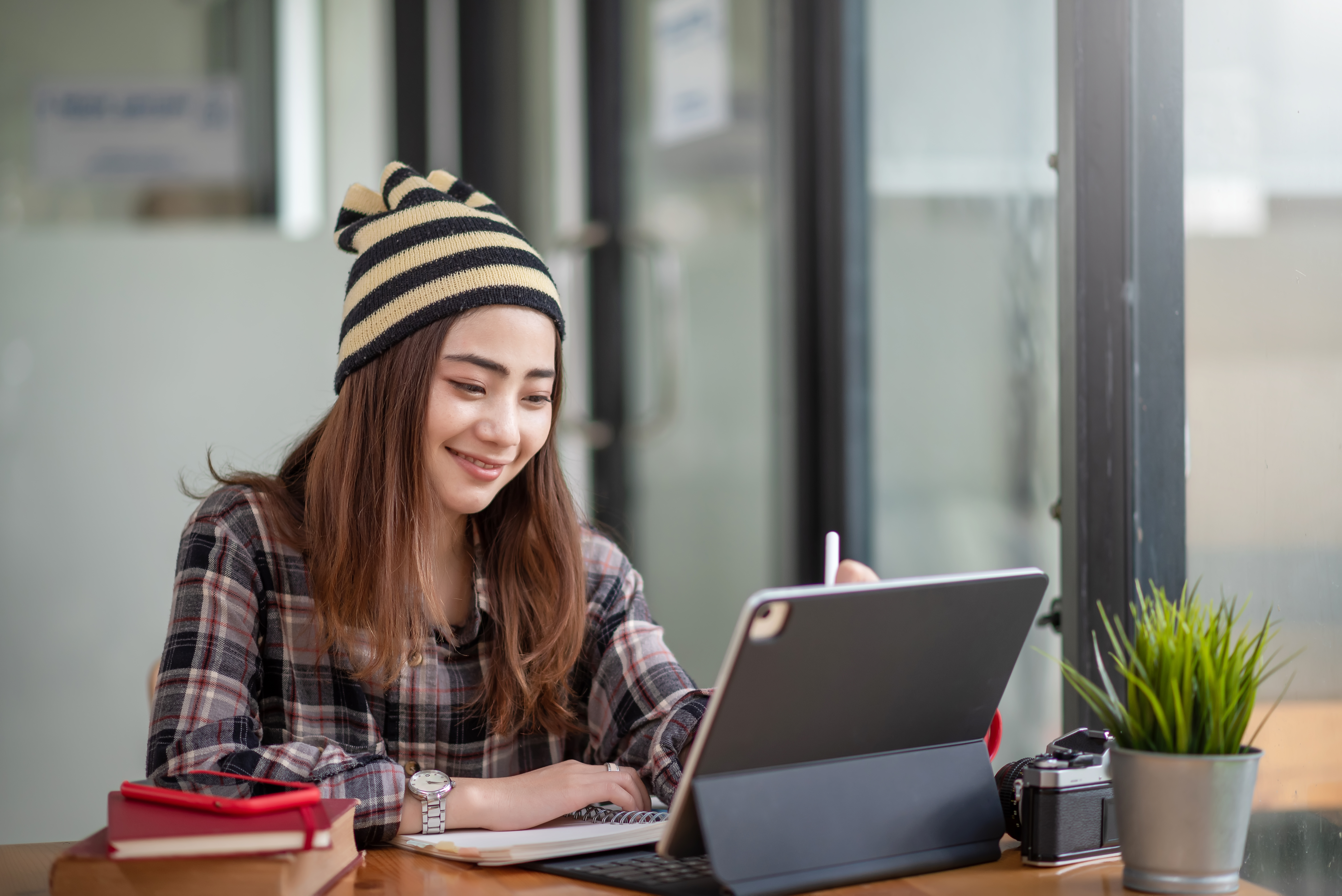 Smiling young woman wearing a striped beanie and plaid shirt works on a laptop or tablet at a wooden desk with a notebook, pen, camera, and small plant nearby. She is looking at the screen and appears to be studying or working remotely in a bright, modern office or study space.