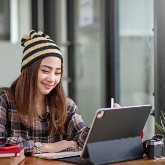 Smiling young woman wearing a striped beanie and plaid shirt works on a laptop or tablet at a wooden desk with a notebook, pen, camera, and small plant nearby. She is looking at the screen and appears to be studying or working remotely in a bright, modern office or study space.