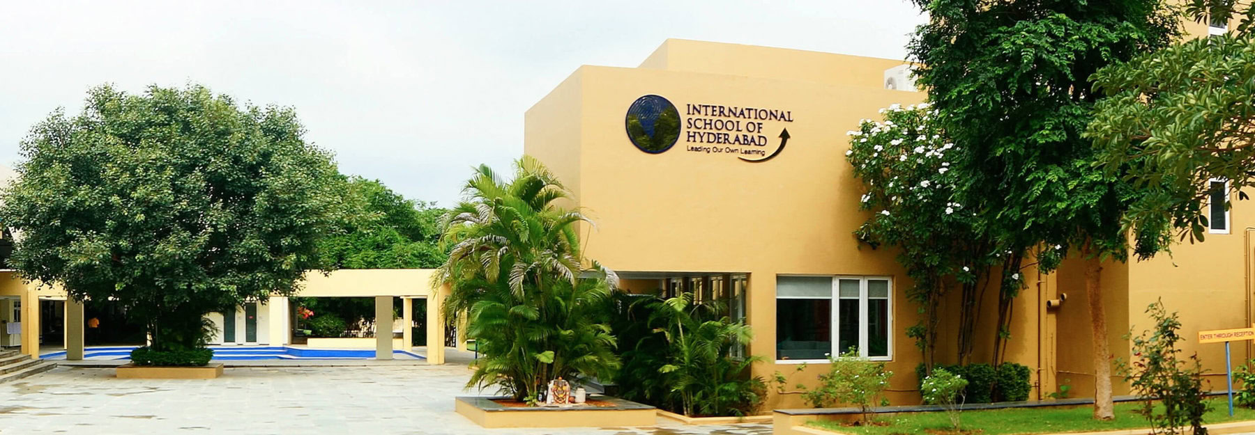 The modern, tan-colored exterior of the International School of Hyderabad, surrounded by lush green trees and tropical plants.