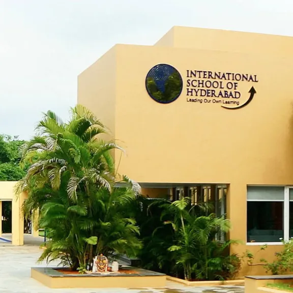 The modern, tan-colored exterior of the International School of Hyderabad, surrounded by lush green trees and tropical plants.