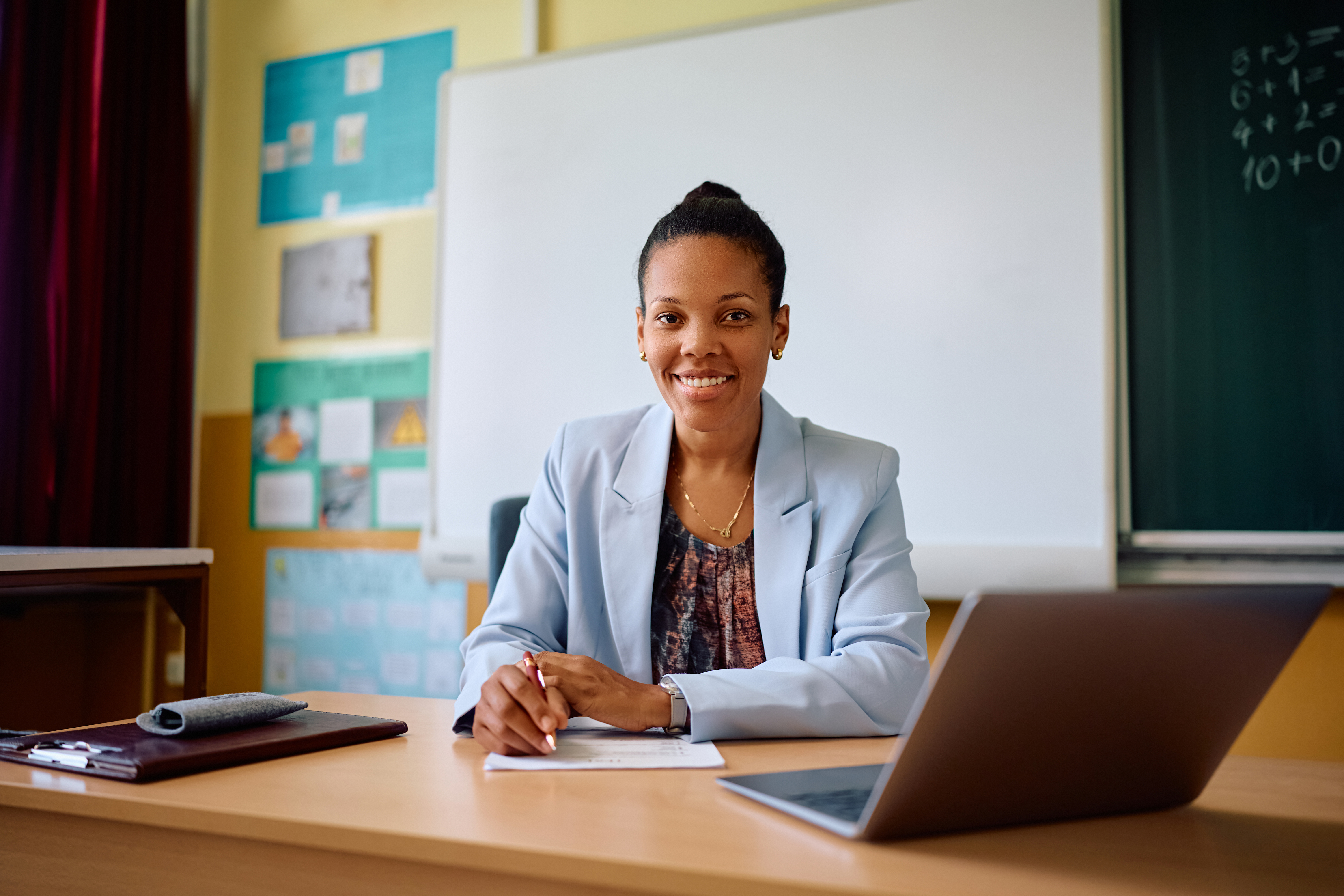 Smiling teacher sitting at a desk with a laptop and paperwork in a classroom setting.