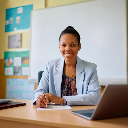 Smiling teacher sitting at a desk with a laptop and paperwork in a classroom setting.