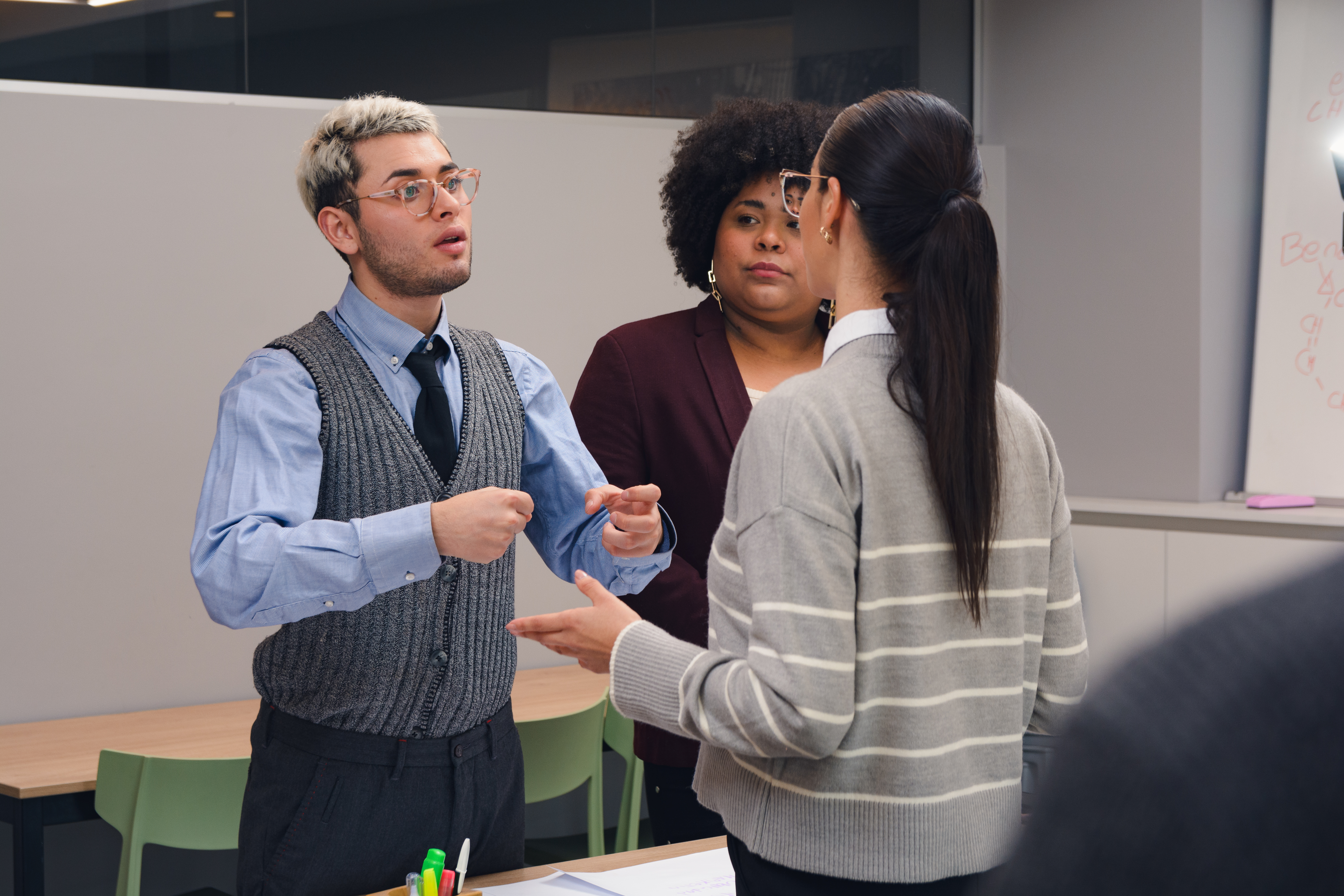 A smiling young woman in a red plaid shirt stands in a modern office, holding a laptop and a coffee cup, representing the flexibility of online study.