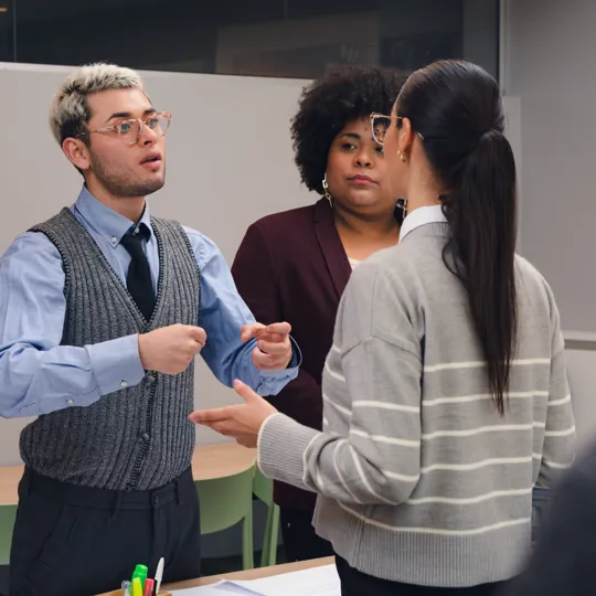 A smiling young woman in a red plaid shirt stands in a modern office, holding a laptop and a coffee cup, representing the flexibility of online study.