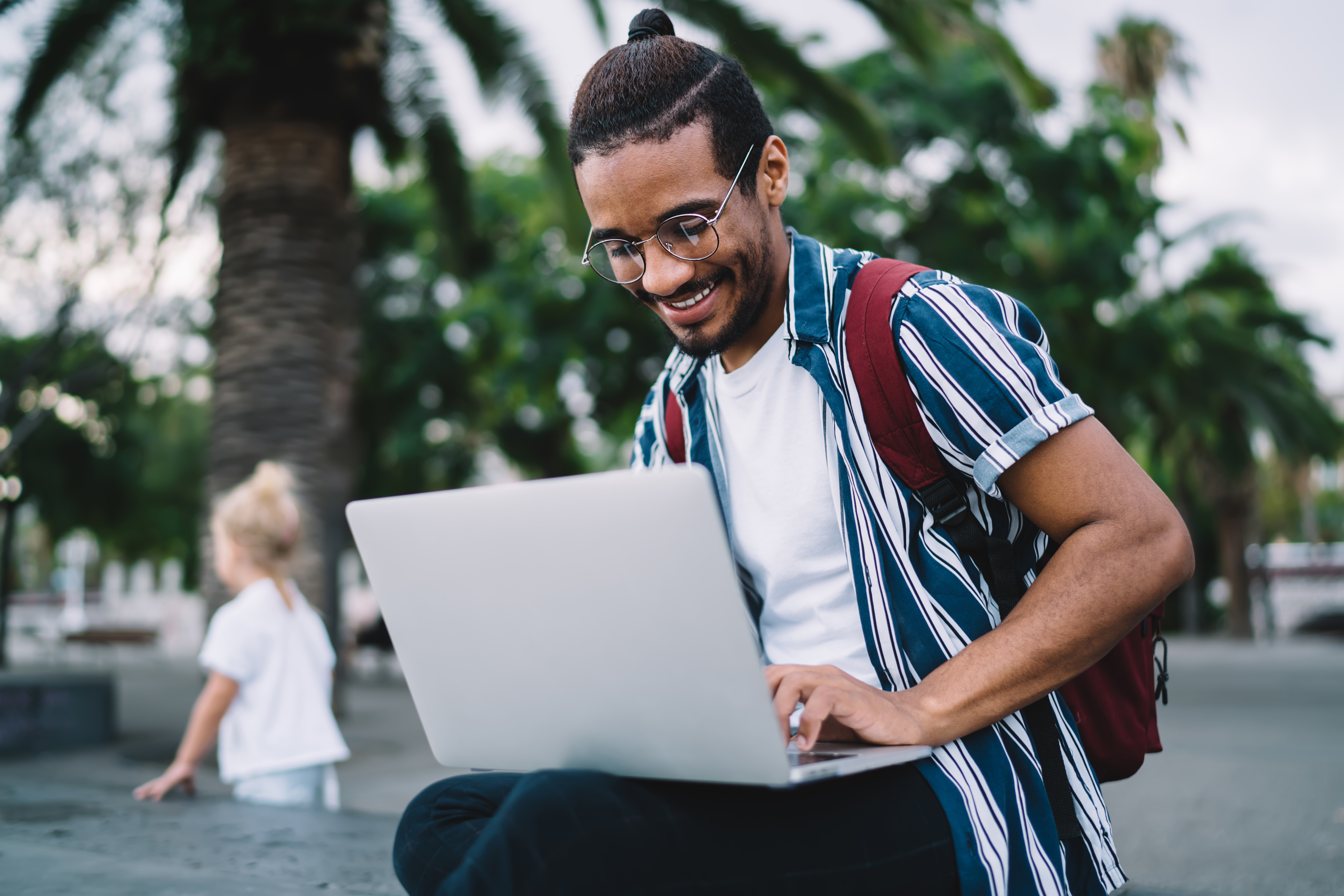 A smiling young man in a striped shirt and glasses works on a laptop while sitting outdoors on a sunny day with palm trees in the background, representing flexible online study.