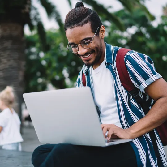 A smiling young man in a striped shirt and glasses works on a laptop while sitting outdoors on a sunny day with palm trees in the background, representing flexible online study.