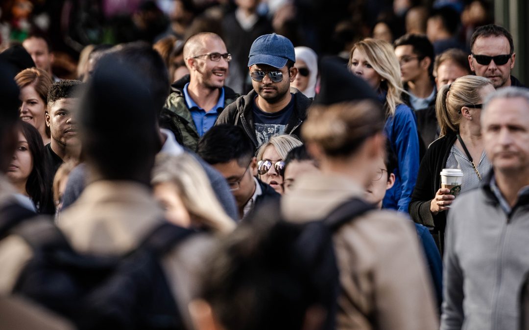  A photograph of a diverse crowd of people in a busy public space, with a man in a blue baseball cap and sunglasses as the focal point.