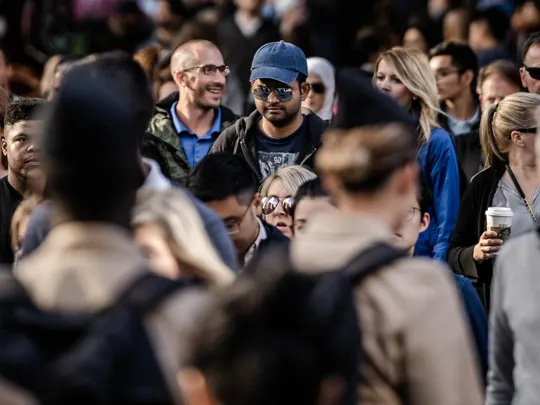 A photograph of a diverse crowd of people in a busy public space, with a man in a blue baseball cap and sunglasses as the focal point.