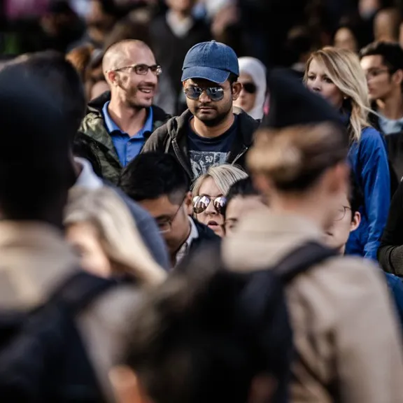 A photograph of a diverse crowd of people in a busy public space, with a man in a blue baseball cap and sunglasses as the focal point.