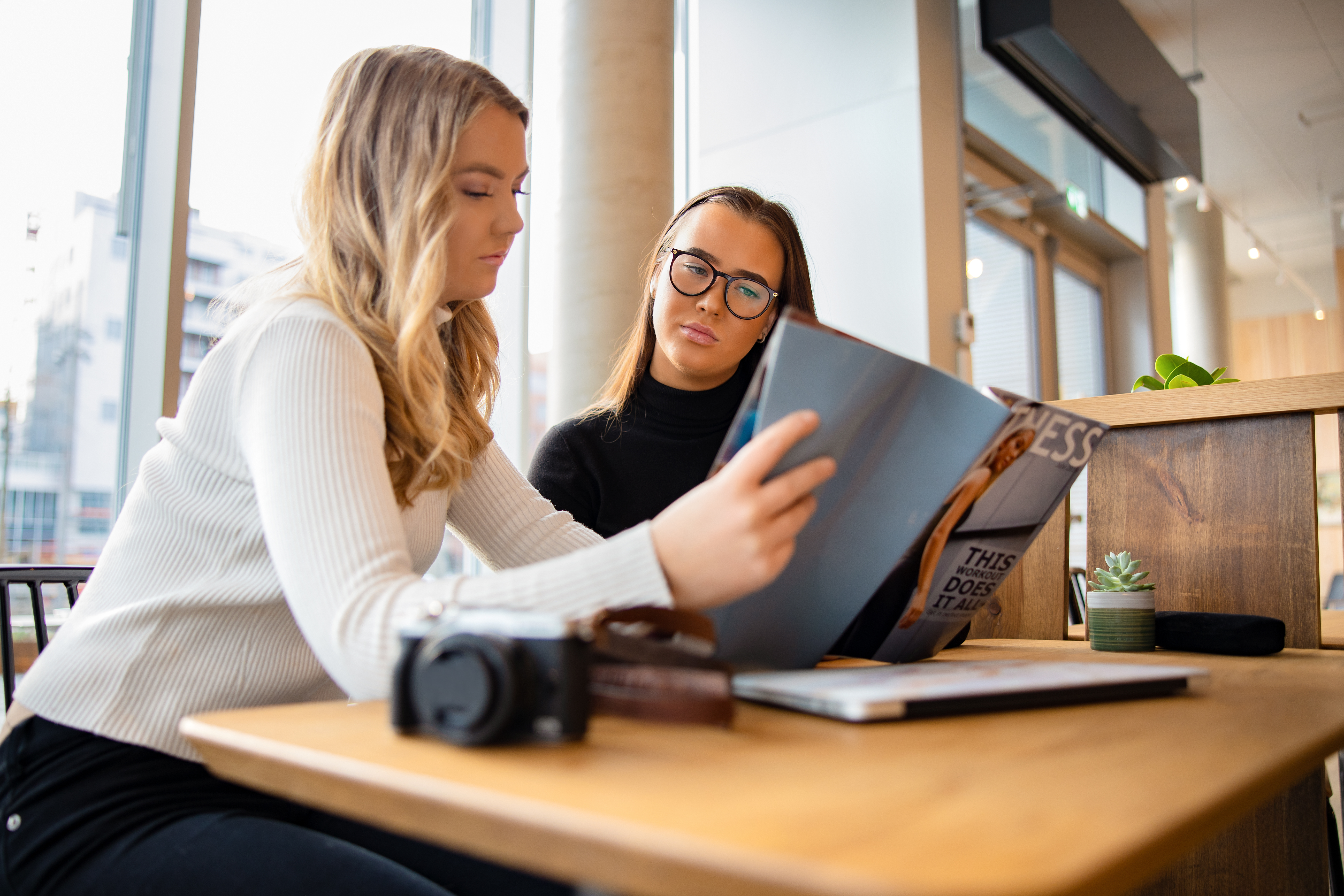 two young women reviewing a magazine together at a wooden table in a bright, modern cafe or co-working space.