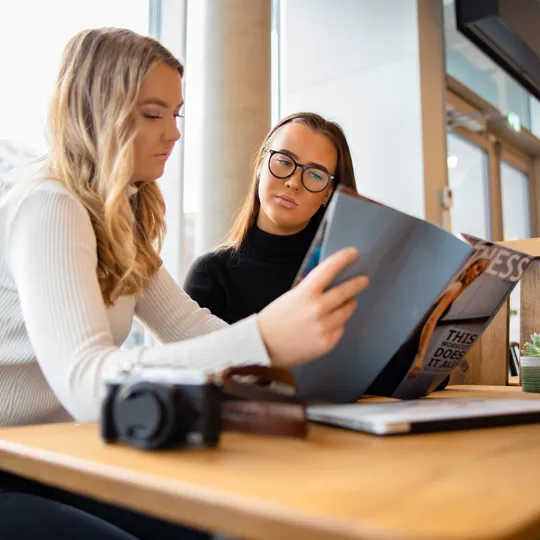 two young women reviewing a magazine together at a wooden table in a bright, modern cafe or co-working space.