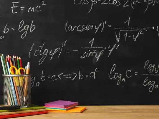 A chalkboard filled with mathematical formulas and equations is behind a wooden desk with a red apple, colorful notebooks, and a cup of writing utensils, symbolizing mathematics applications.