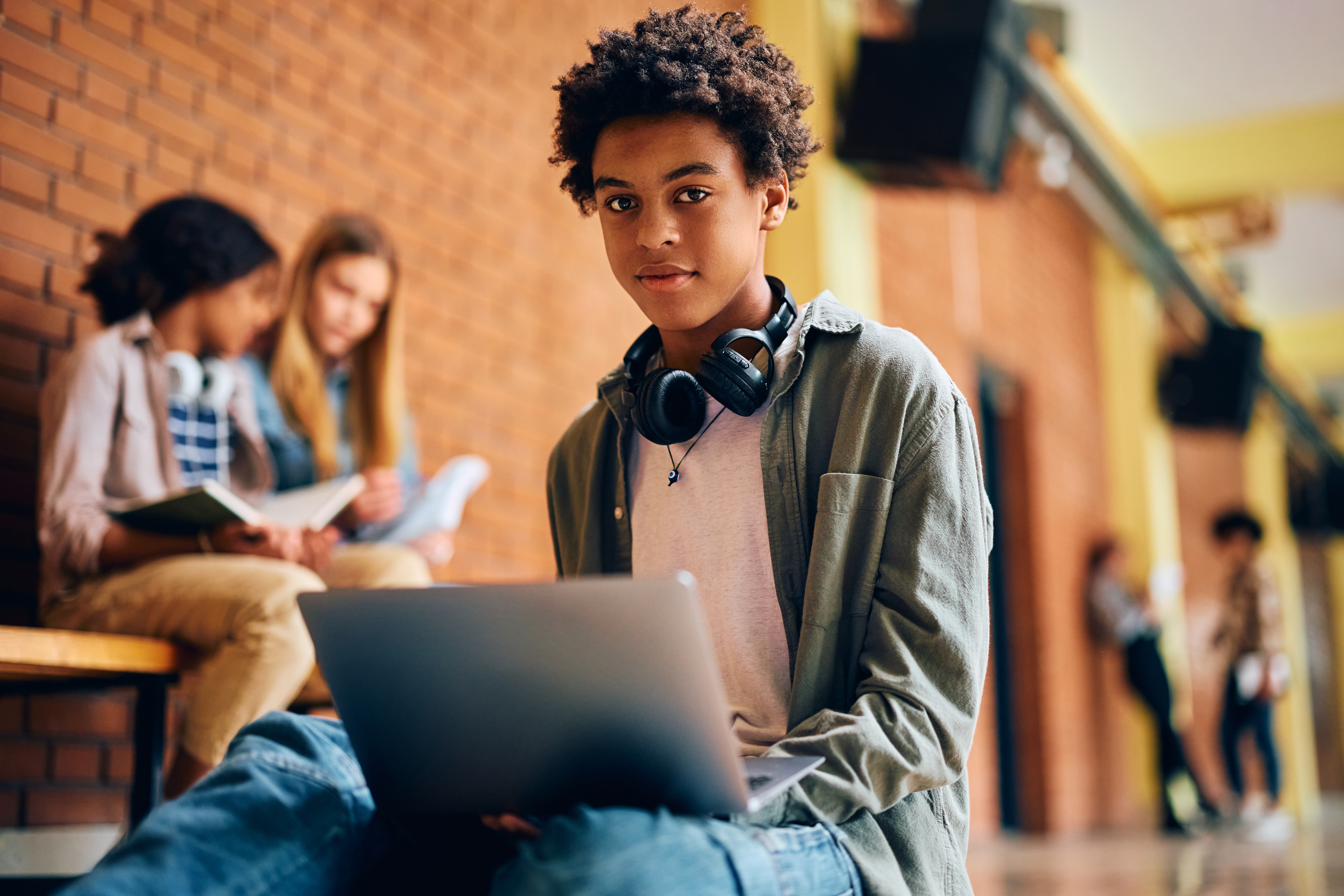 A male high school or university student with headphones around his neck working on a laptop in a school hallway with other students in the background.
