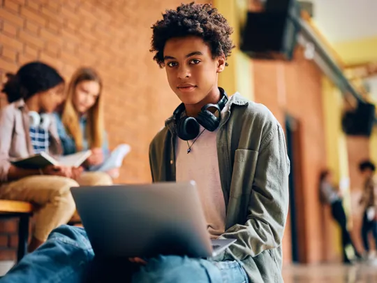 A male high school or university student with headphones around his neck working on a laptop in a school hallway with other students in the background.
