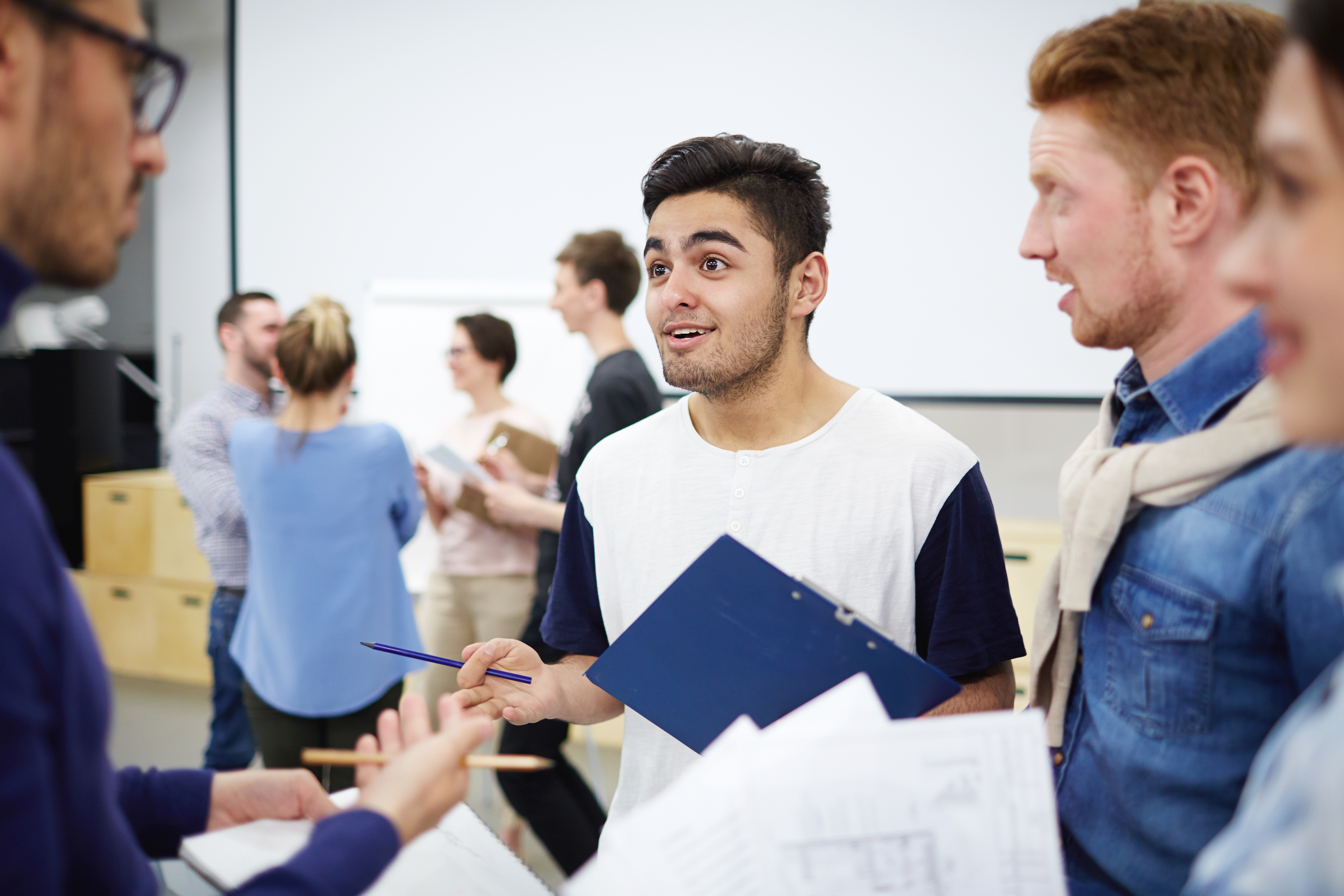 A young man holding a clipboard smiles and gestures with a pencil while engaging in an excited discussion with a small group of classmates in a modern classroom setting.