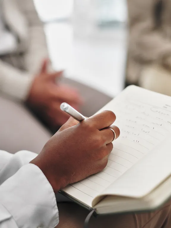 A close-up of a person's hands writing notes in an open notebook with a silver pen, with two blurred people sitting in the background, symbolizing psychology or counseling.