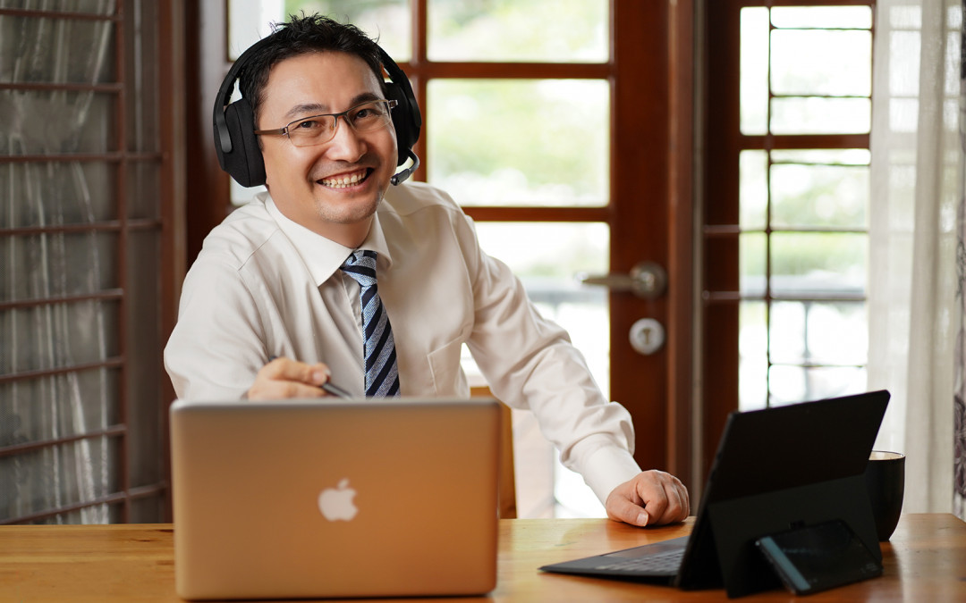 A friendly, smiling man wearing glasses, a shirt, and a tie, sits at a desk with two laptops, wearing an over-ear headset with a microphone. He appears to be an online instructor, tutor, or professional working remotely in a well-lit office or home setting.