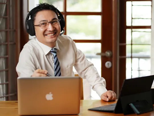 A friendly, smiling man wearing glasses, a shirt, and a tie, sits at a desk with two laptops, wearing an over-ear headset with a microphone. He appears to be an online instructor, tutor, or professional working remotely in a well-lit office or home setting.