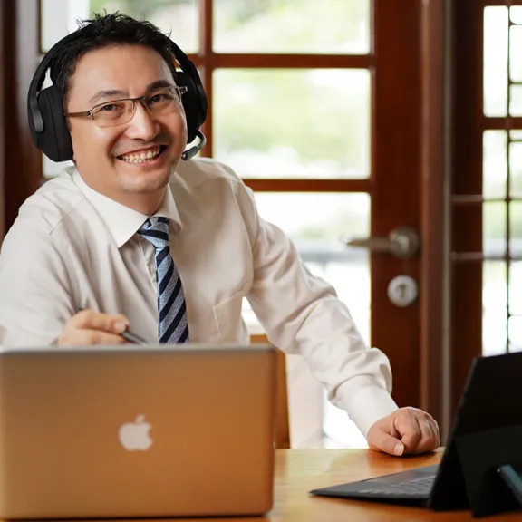 A friendly, smiling man wearing glasses, a shirt, and a tie, sits at a desk with two laptops, wearing an over-ear headset with a microphone. He appears to be an online instructor, tutor, or professional working remotely in a well-lit office or home setting.