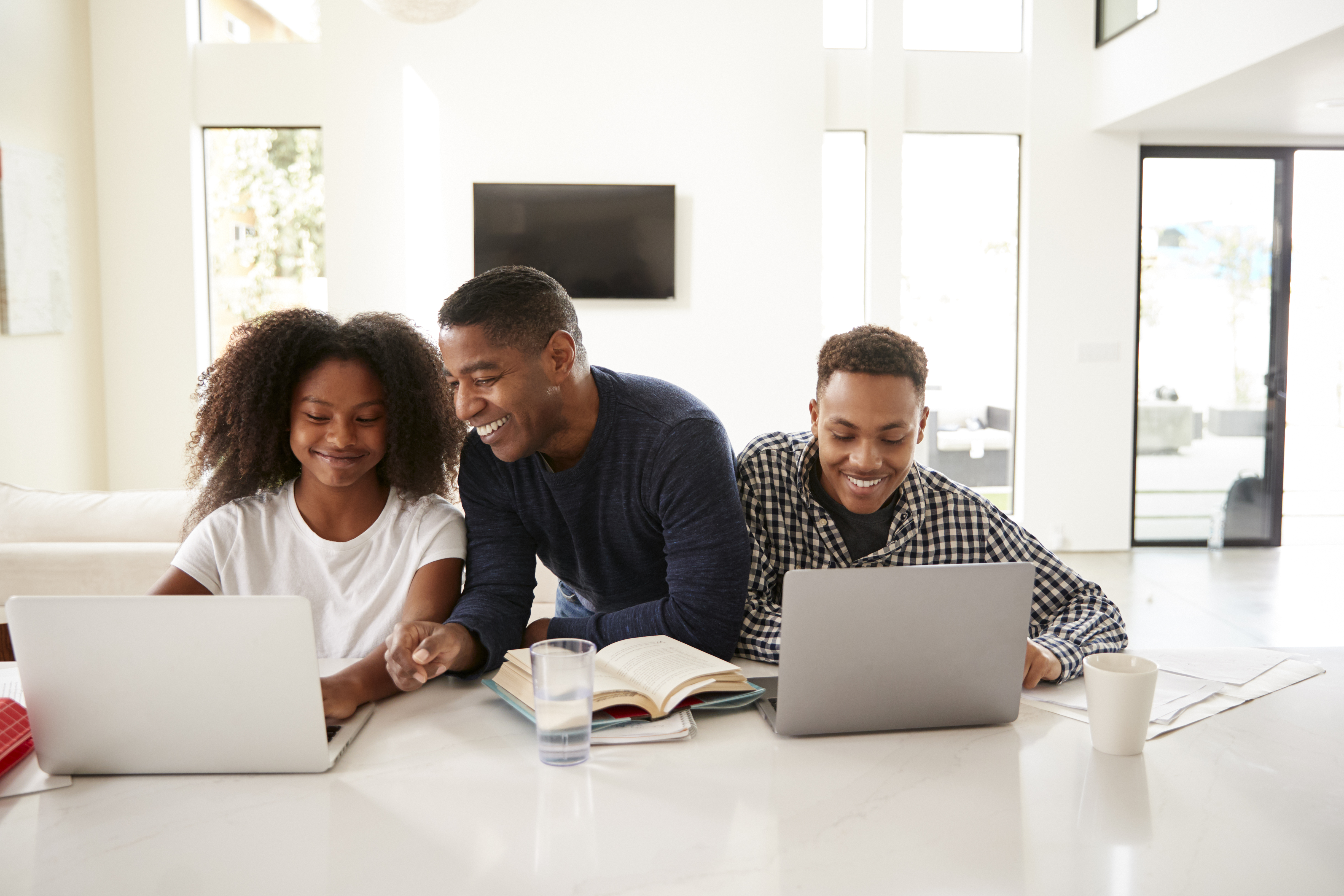 Smiling international family with a father supporting his two children as they study online with laptops at home. This image represents Pamoja Education providing consistent and flexible academic structure for students in international families, fostering success and engagement across different locations and time zones.