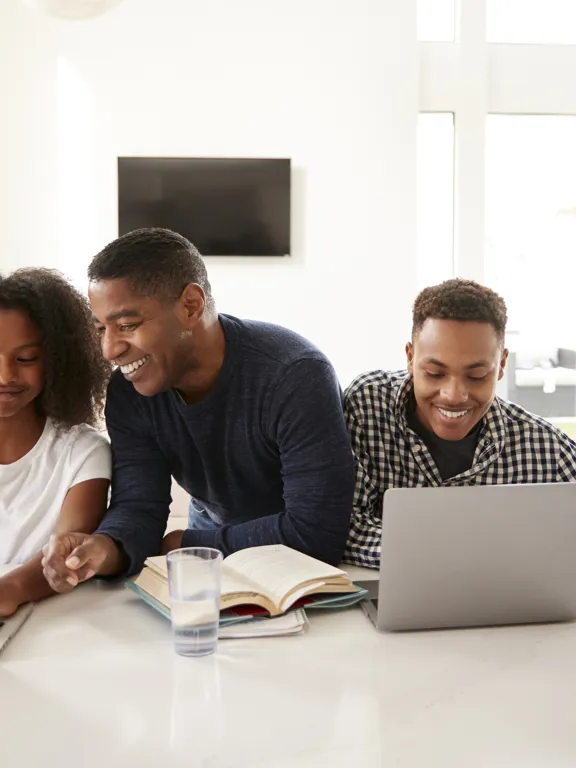 Smiling international family with a father supporting his two children as they study online with laptops at home. This image represents Pamoja Education providing consistent and flexible academic structure for students in international families, fostering success and engagement across different locations and time zones.