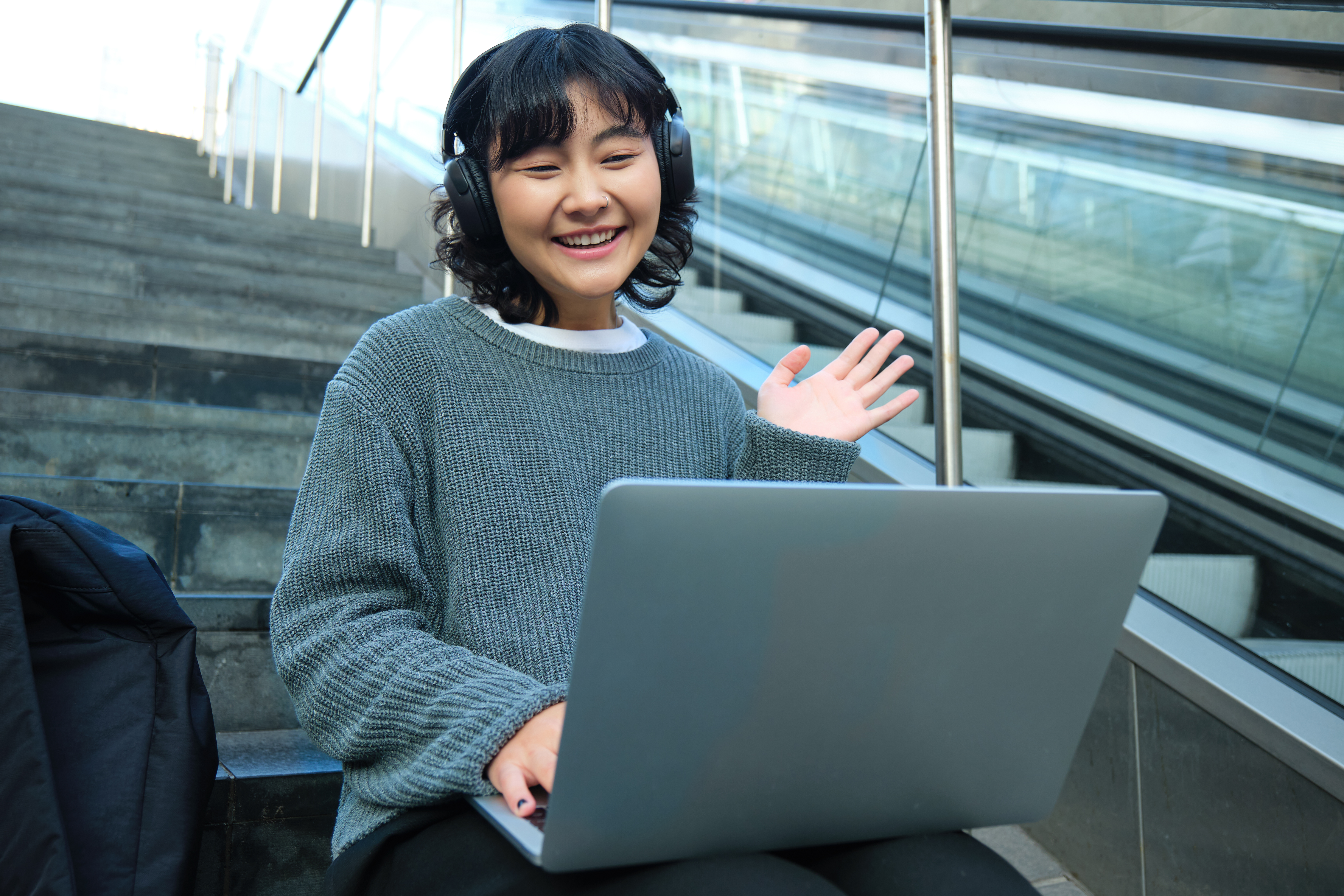 A young woman in a grey sweater and black headphones smiles and waves while working on a laptop on outdoor steps, representing flexible online learning.