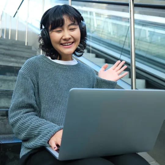 A young woman in a grey sweater and black headphones smiles and waves while working on a laptop on outdoor steps, representing flexible online learning.