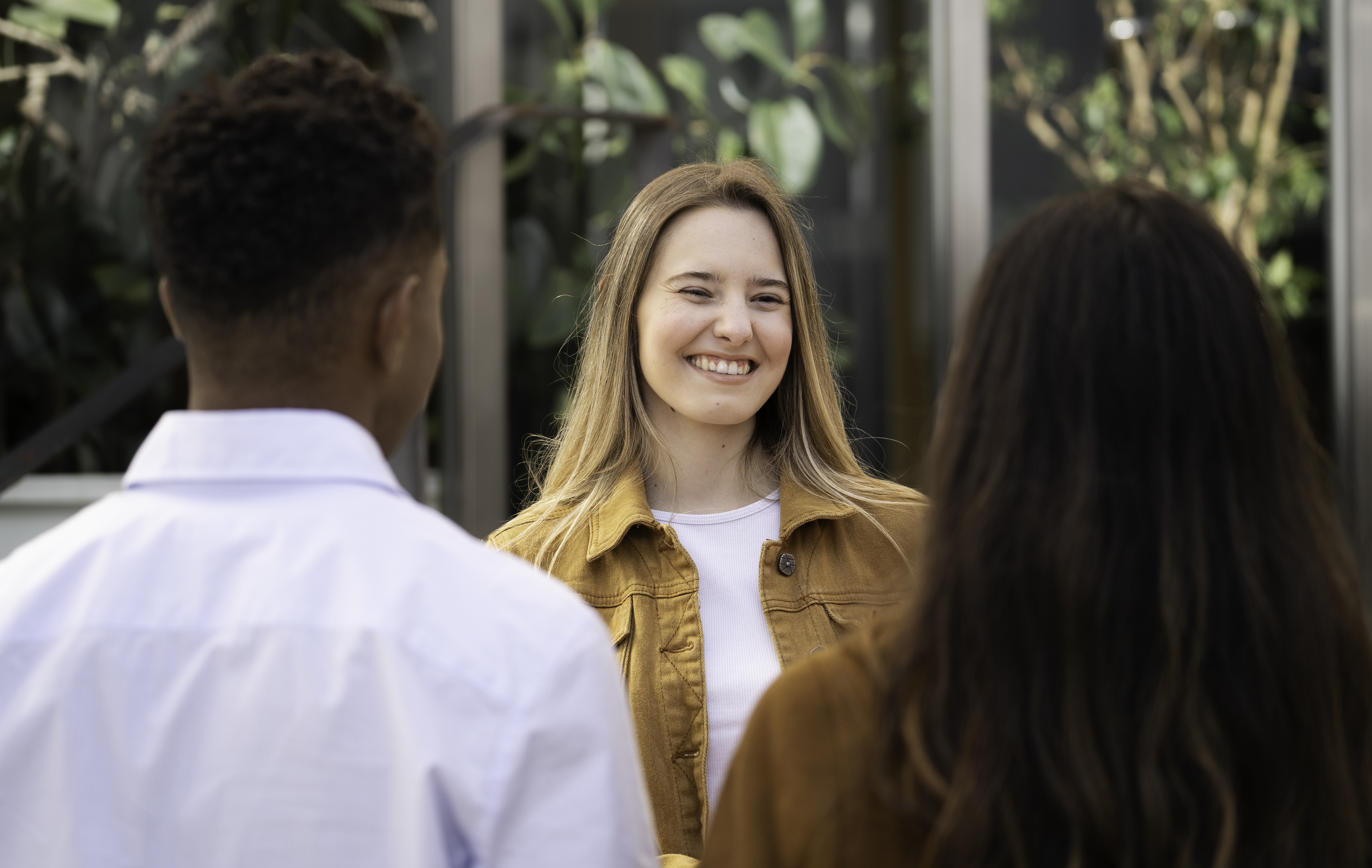 A smiling young woman with blonde hair and black headphones waves during a video call on her laptop while sitting on outdoor steps, representing flexible online computer science study.