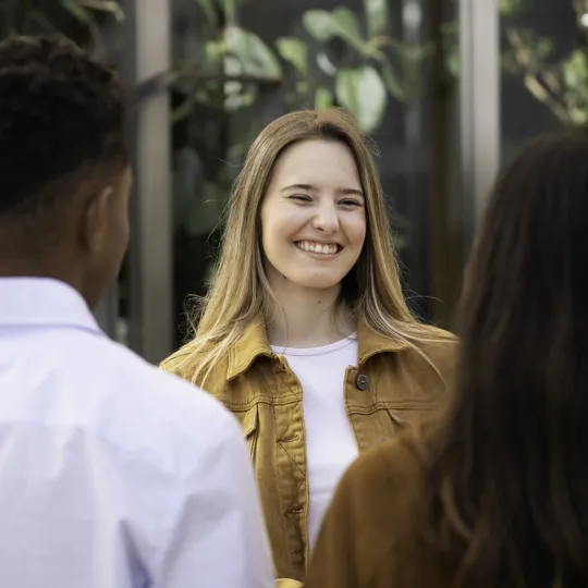 A smiling young woman with blonde hair and black headphones waves during a video call on her laptop while sitting on outdoor steps, representing flexible online computer science study.