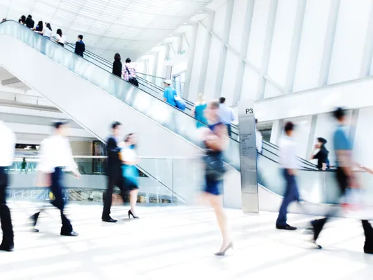 A young woman in a plaid shirt smiles and gestures enthusiastically while talking to a person in an outdoor cafe setting about business management.