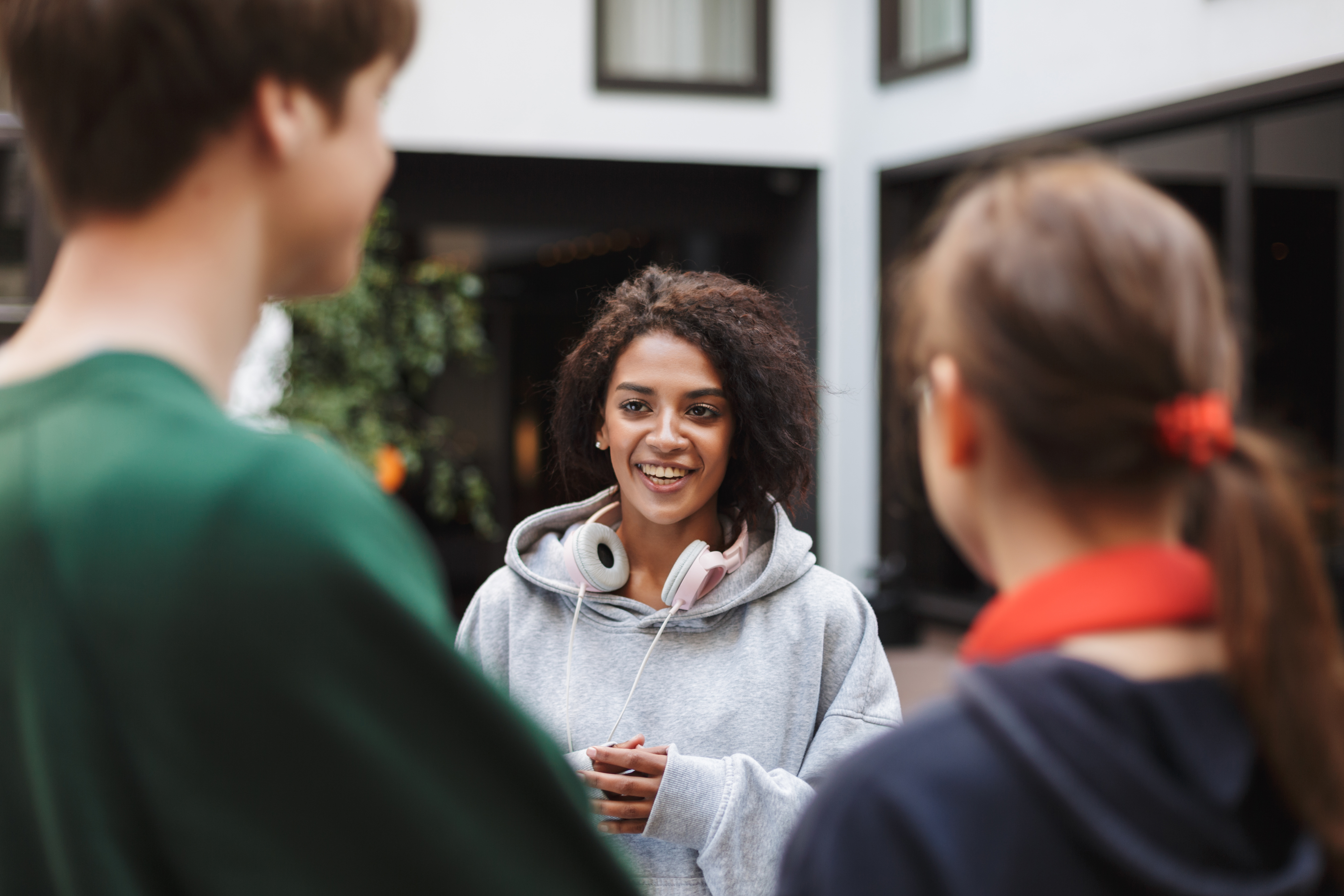 A smiling student with curly hair in a grey hoodie and pink headphones, talking with two classmates in a school courtyard.
