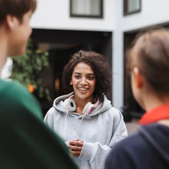 A smiling student with curly hair in a grey hoodie and pink headphones, talking with two classmates in a school courtyard.
