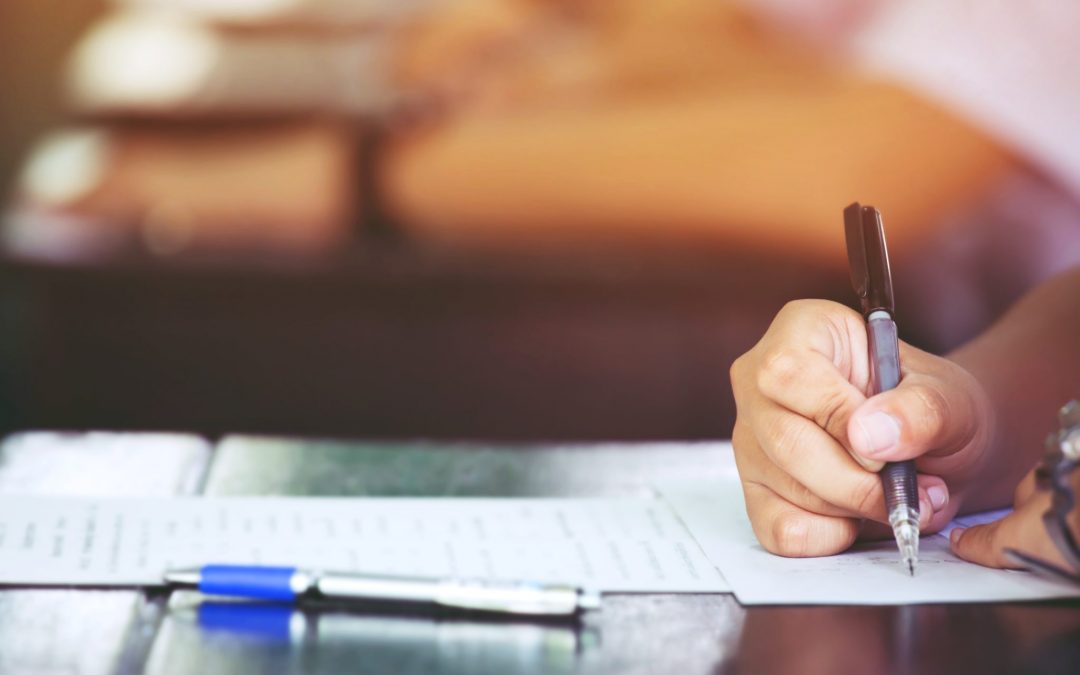 Close-up of a hand holding a black pen and writing on a paper or test sheet on a desk, with a blue pen lying nearby. The background is blurred with warm, soft lighting, suggesting a classroom or exam setting.