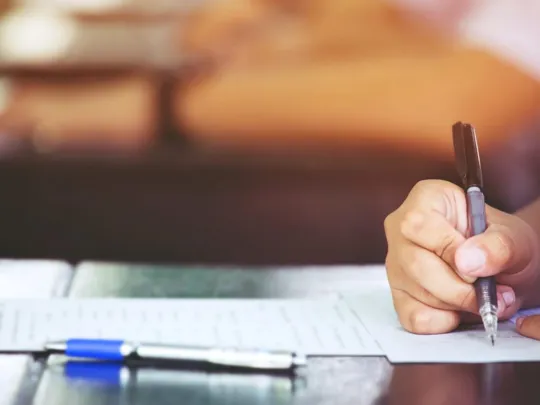 Close-up of a hand holding a black pen and writing on a paper or test sheet on a desk, with a blue pen lying nearby. The background is blurred with warm, soft lighting, suggesting a classroom or exam setting.