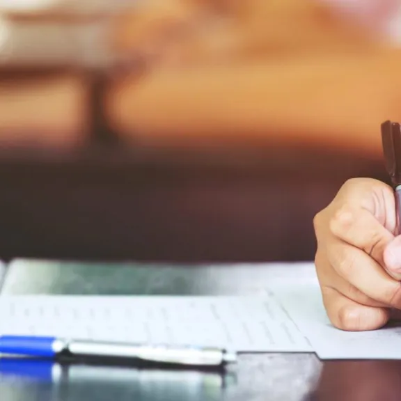 Close-up of a hand holding a black pen and writing on a paper or test sheet on a desk, with a blue pen lying nearby. The background is blurred with warm, soft lighting, suggesting a classroom or exam setting.