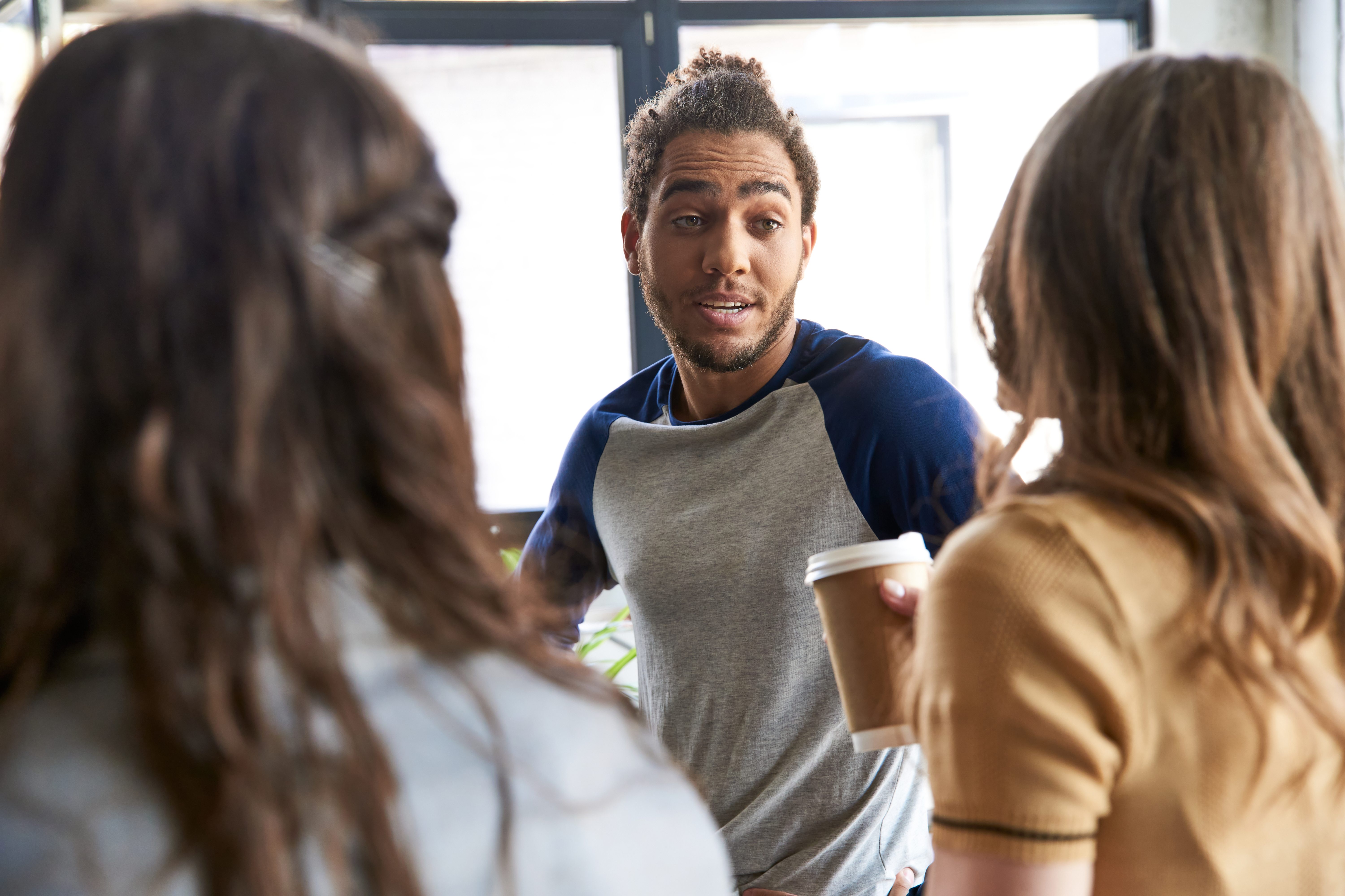 A young man holding a coffee cup and talking to two women in an indoor setting with a large window in the background.