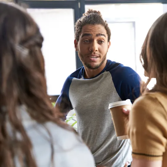 A young man holding a coffee cup and talking to two women in an indoor setting with a large window in the background.